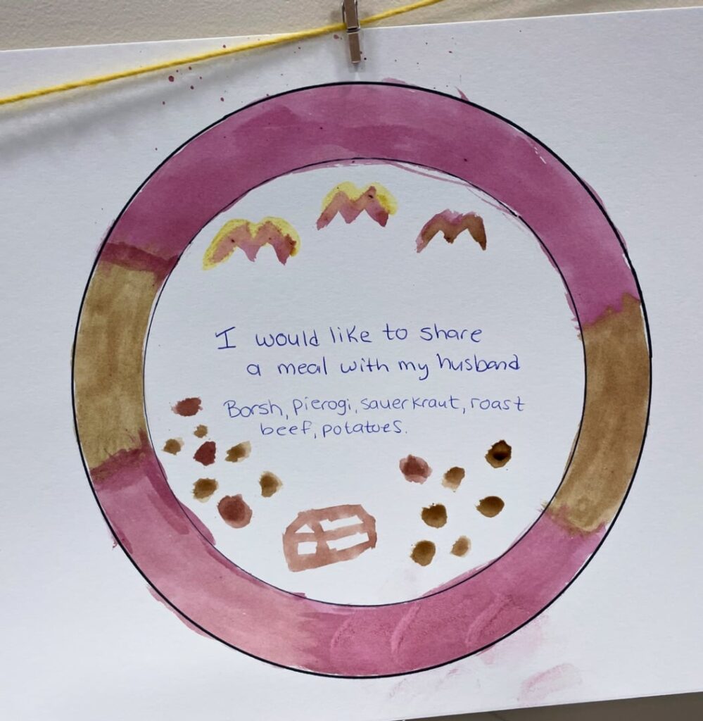A painting of a plate with food on it. A written message on the plate reads "I would like to share a meal with my husband. Borsh, pierogi, saurkraut, roast, beef, potatoes." 
